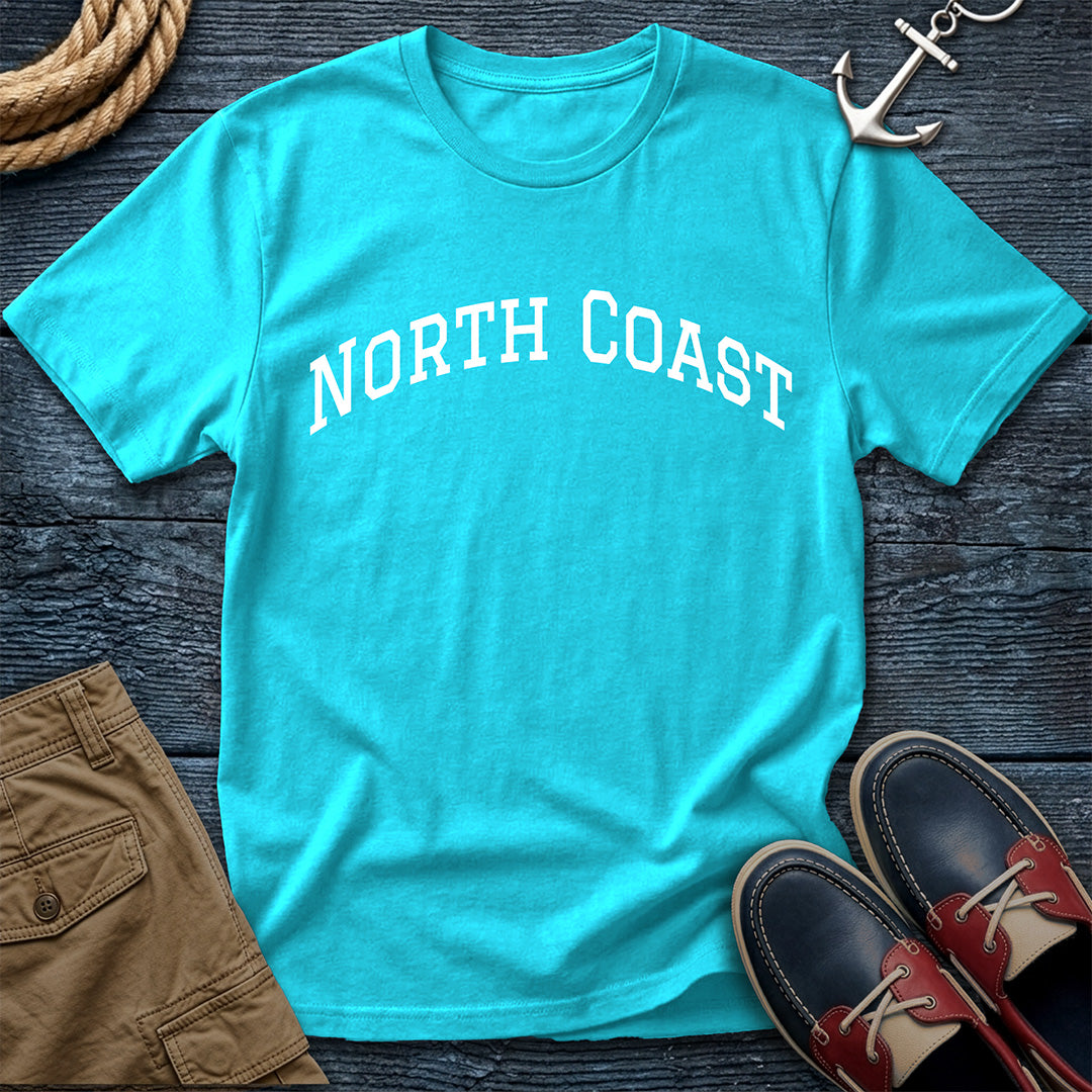 A turquoise crewneck t-shirt with 'NORTH COAST' printed in white arched text across the chest, styled in a flat lay on a dark wood background with an anchor, boat shoes, shorts, and rope.