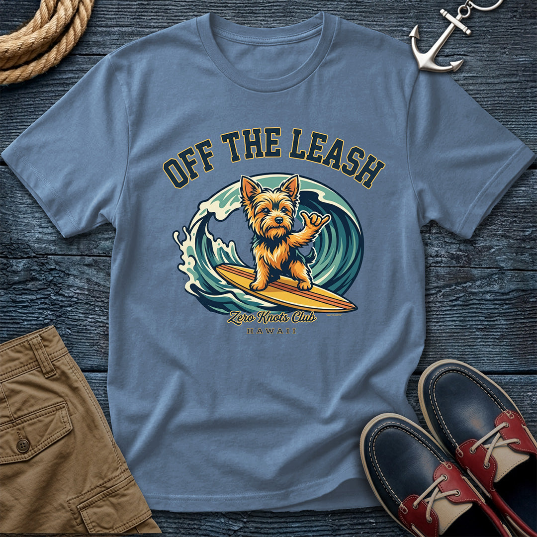 Blue t-shirt with a dog surfing graphic and text on a wooden surface with shoes and shorts.