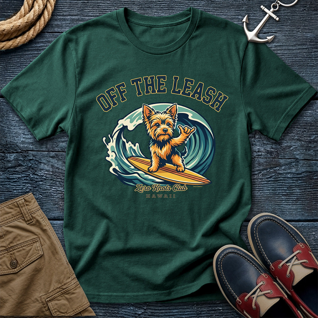 Green t-shirt with a dog surfing graphic on a wooden surface with shoes and shorts.
