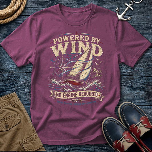 A berry short-sleeve graphic t-shirt laid flat on a dark wood background, styled with a rope, silver anchor, tan shorts, and navy-and-red boat shoes. The shirt features a vintage-style sailboat and wave graphic with the text 'POWERED BY WIND' and 'NO ENGINE REQUIRED'.