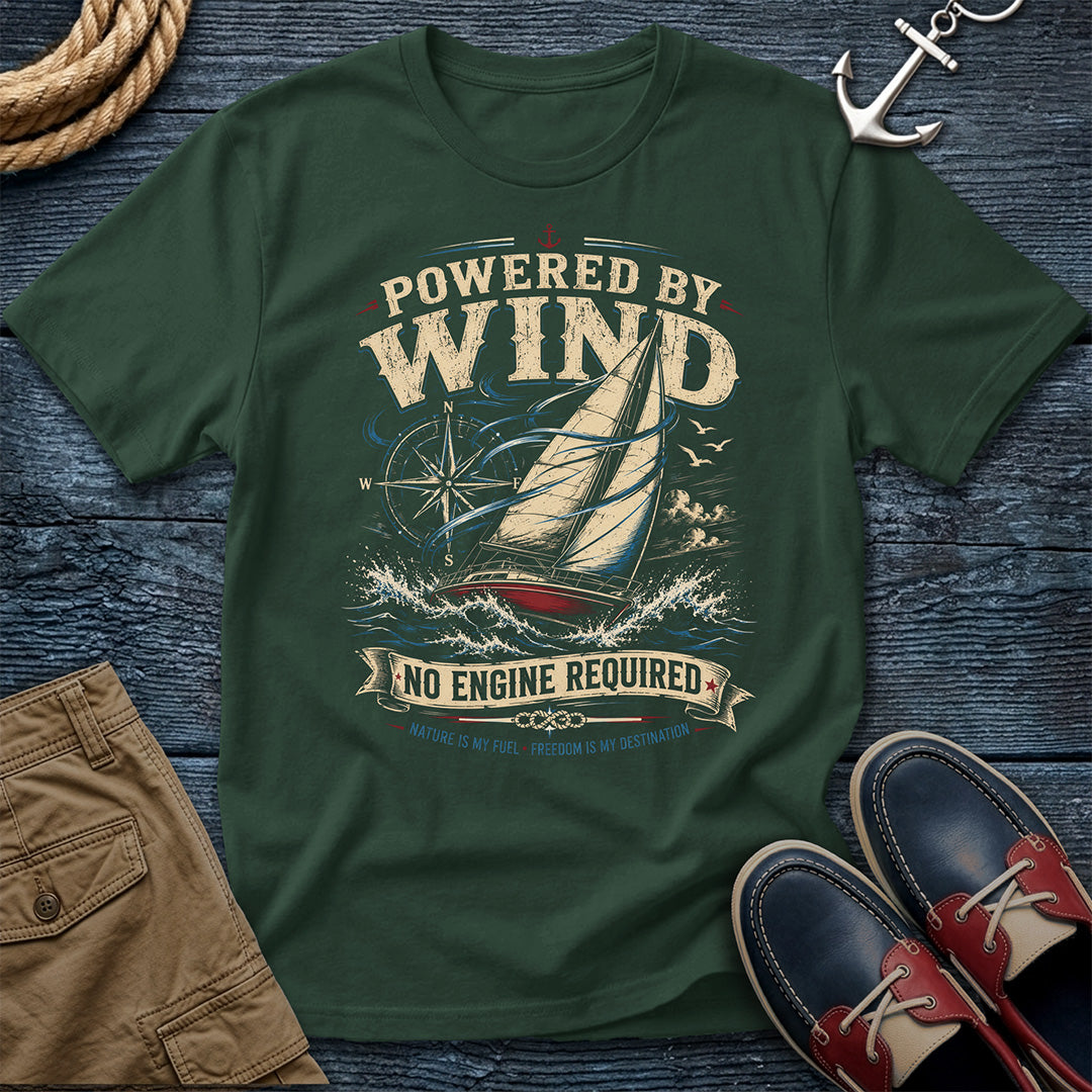 A dark green crewneck t-shirt featuring a vintage-style sailboat graphic with the text 'POWERED BY WIND' and 'NO ENGINE REQUIRED'. The shirt is displayed in a flat-lay arrangement on a dark blue wooden background, styled with a rope, an anchor, khaki shorts, and red-and-navy boat shoes.