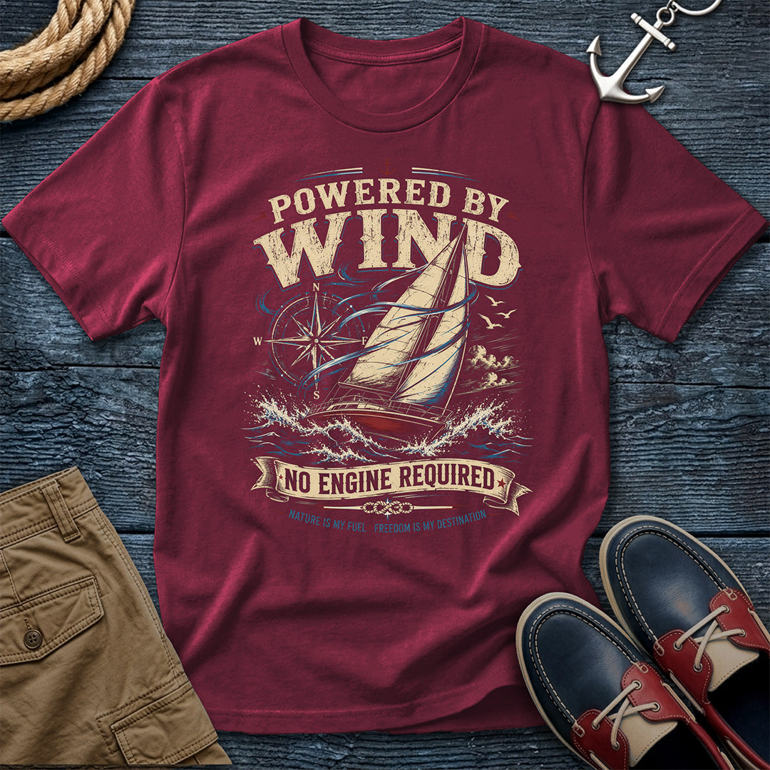 A maroon short-sleeve graphic t-shirt laid flat on a dark wood background, surrounded by nautical and casual props including a rope, anchor, khaki shorts, and navy-and-red boat shoes. The shirt features a large vintage-style sailboat graphic with the text 'POWERED BY WIND' and 'NO ENGINE REQUIRED'.