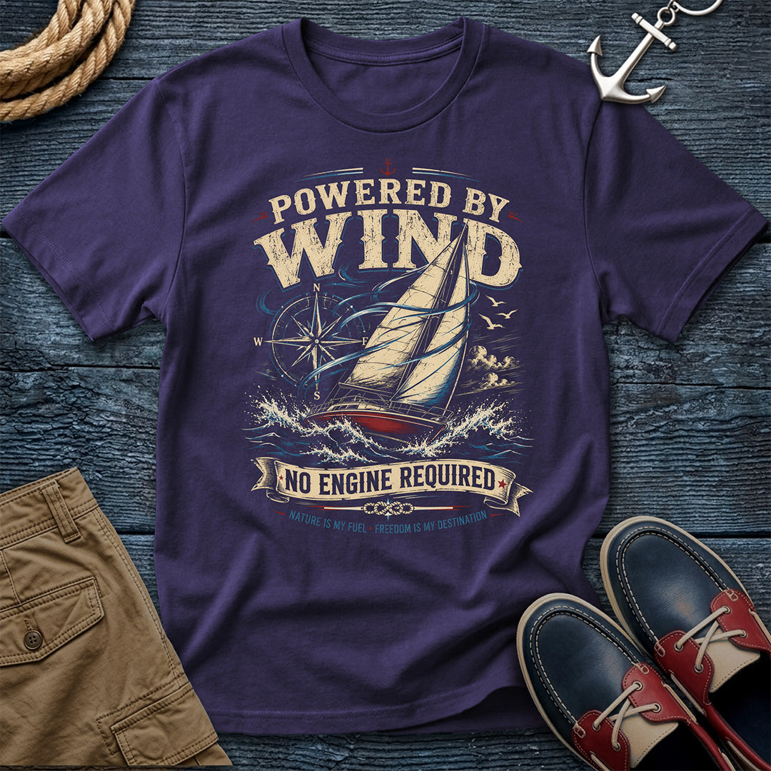 A Grape colored graphic t-shirt featuring a sailboat and the text 'POWERED BY WIND NO ENGINE REQUIRED' in a vintage nautical design. The shirt is styled in a flat-lay arrangement on a dark wood background with a rope, khaki shorts, an anchor, and red-and-navy boat shoes.