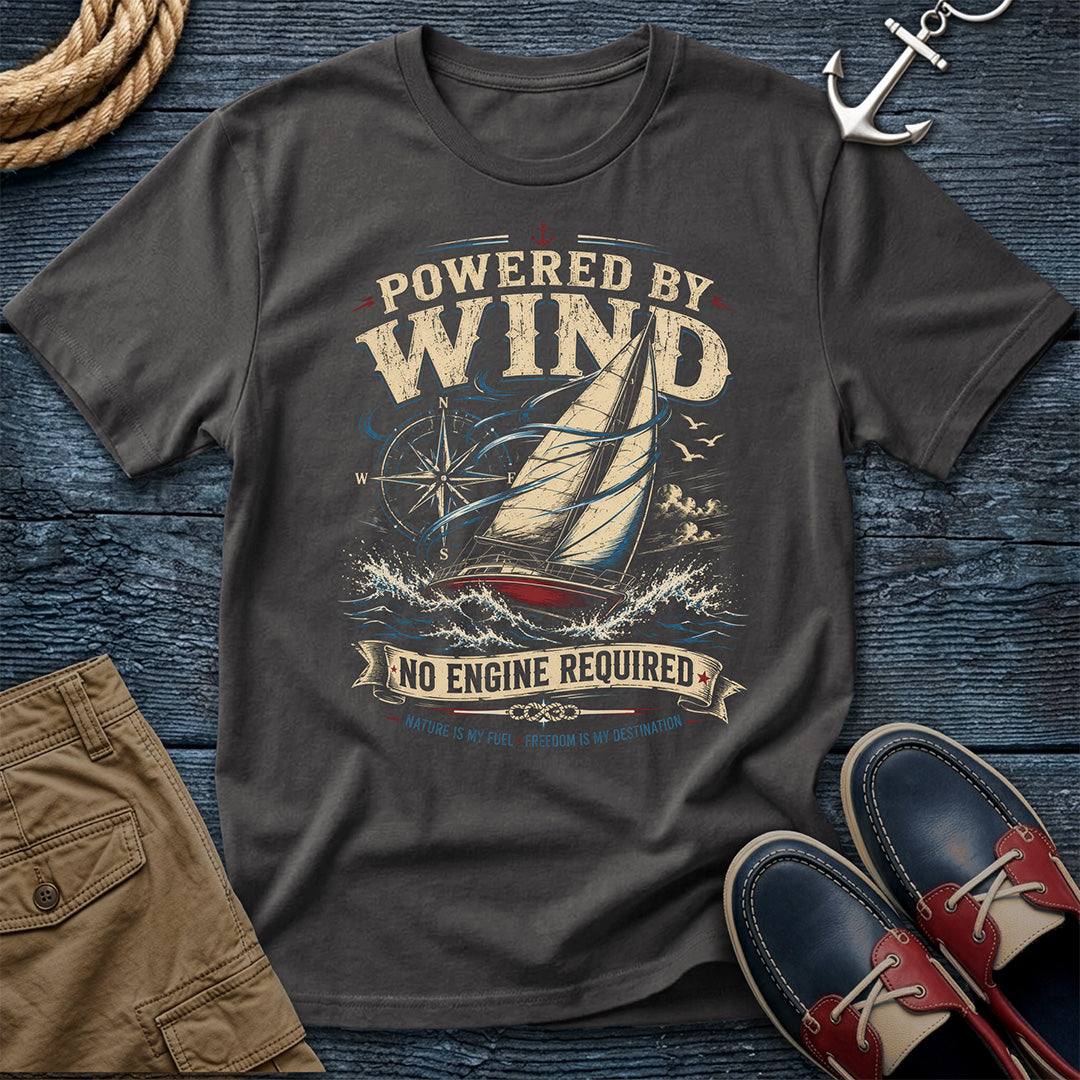 A graphite colored graphic t-shirt featuring a sailboat and the text 'POWERED BY WIND NO ENGINE REQUIRED' in a vintage nautical design. The shirt is styled in a flat-lay arrangement on a dark wood background with a rope, khaki shorts, an anchor, and red-and-navy boat shoes.