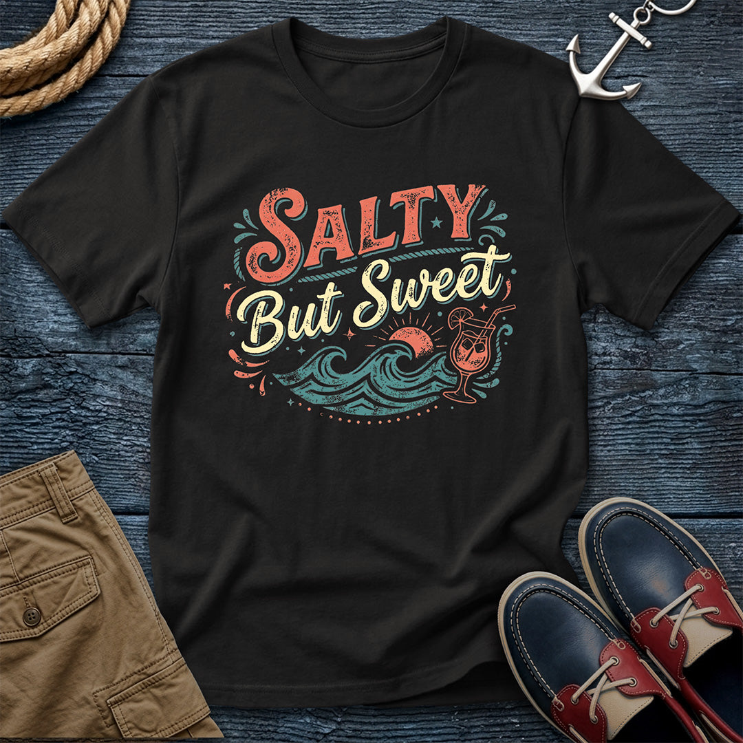 Black t-shirt with 'Salty But Sweet' design on a wooden surface with shorts and shoes.