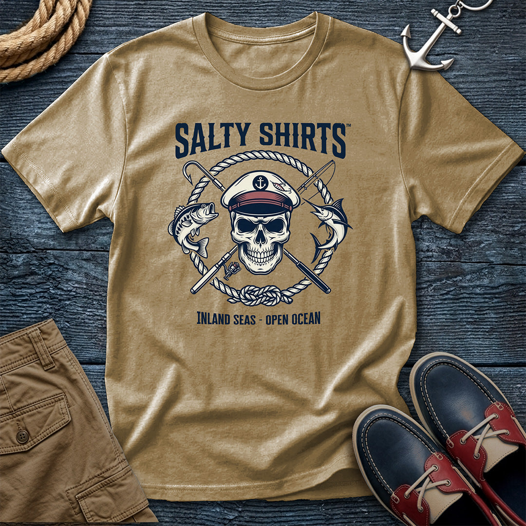 Khaki t-shirt with skull and crossed swords design on a wooden surface with shoes and shorts.
