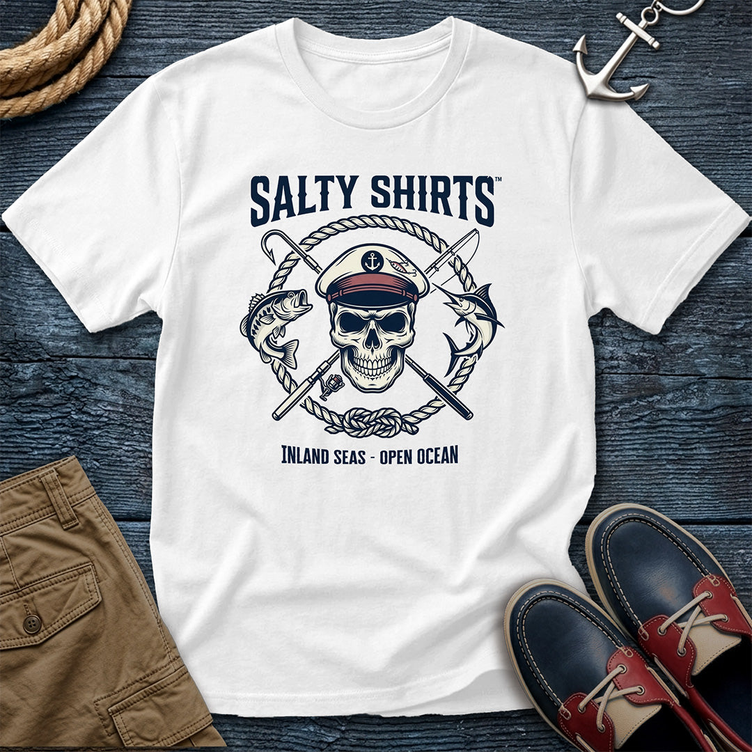 White t-shirt with skull and crossed swords design on a wooden surface with shoes and shorts.