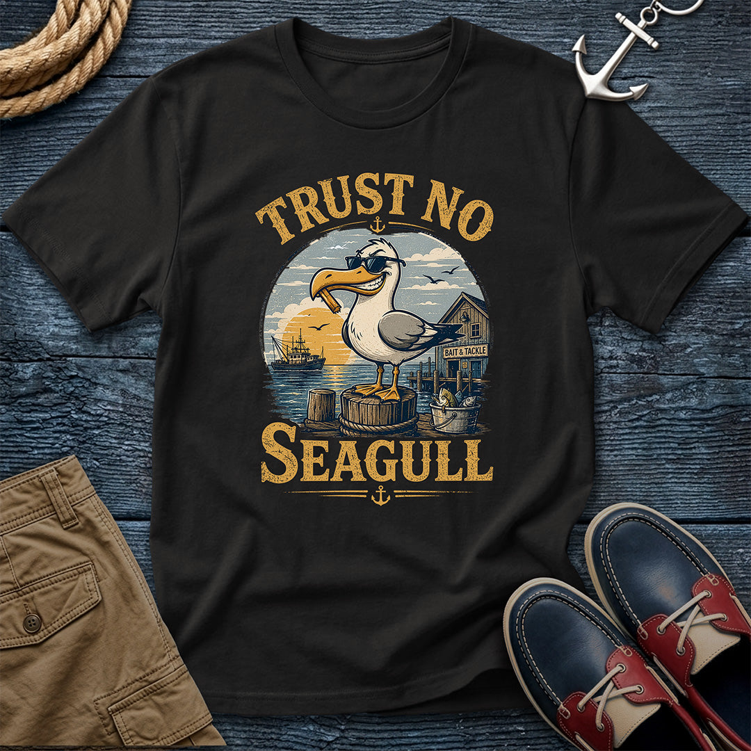 A black graphic t-shirt featuring a vintage-style illustration of a seagull by the harbor with the text 'TRUST NO SEAGULL'. The shirt is presented in a flat-lay arrangement on a dark blue wooden background, styled with a rope, khaki shorts, a silver anchor, and red-and-navy boat shoes.