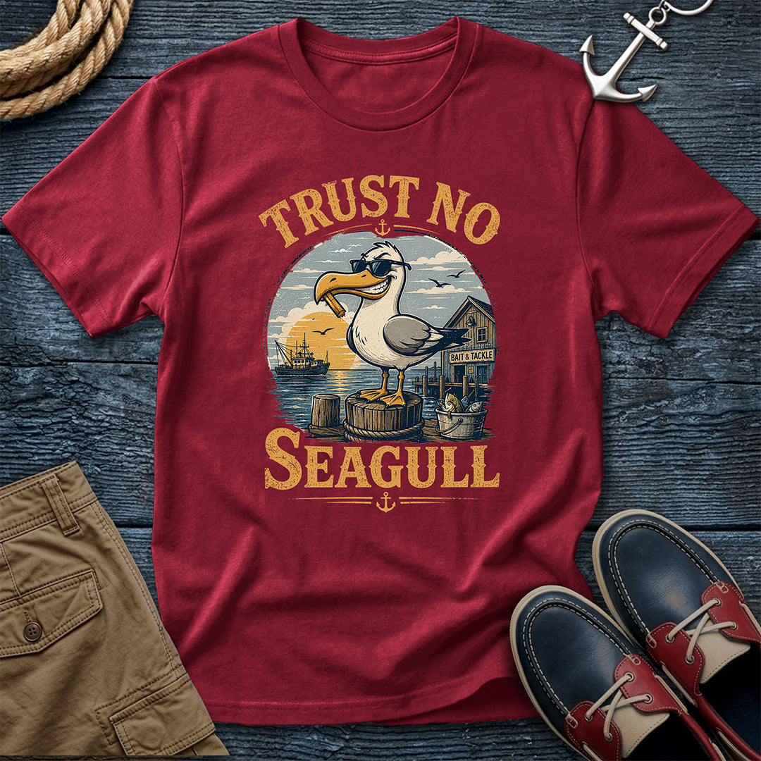 A red short-sleeve graphic T-shirt laid flat on a dark blue wood surface, surrounded by nautical-themed props including a rope, anchor, khaki shorts, and navy-and-red boat shoes. The shirt graphic shows a seagull standing on a dock with the text 'TRUST NO SEAGULL' in large yellow letters.