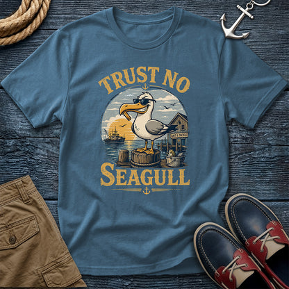 A blue crewneck t-shirt with a vintage-style seagull graphic and the text 'TRUST NO SEAGULL' in yellow. The shirt is styled in a flat lay on a dark wood background with a rope, khaki shorts, a silver anchor, and red-and-navy boat shoes.
