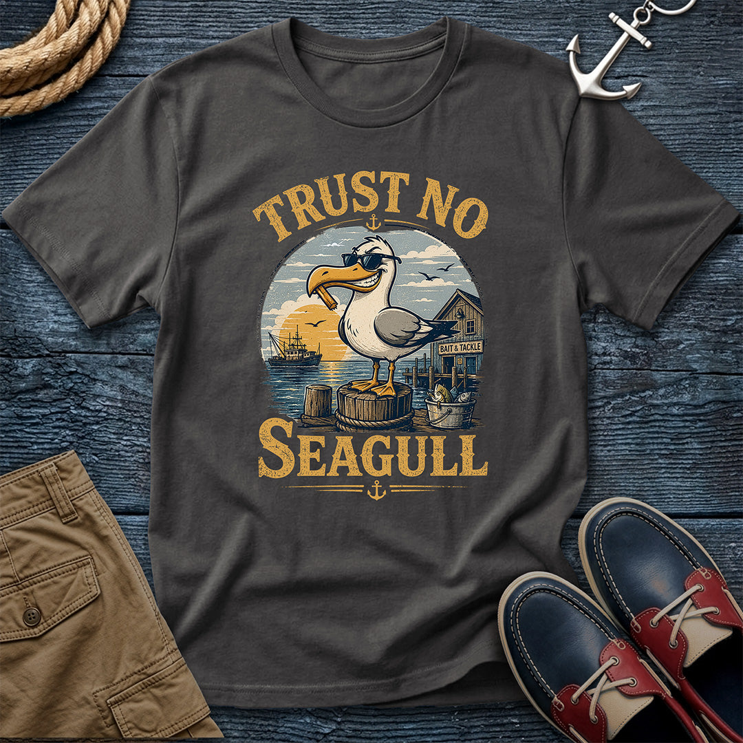 A dark charcoal gray t-shirt with a vintage-style nautical graphic of a seagull and the text 'TRUST NO SEAGULL'. The shirt is presented in a flat-lay arrangement on a dark blue wood background, styled with a rope, khaki shorts, a silver anchor, and red-and-navy boat shoes.