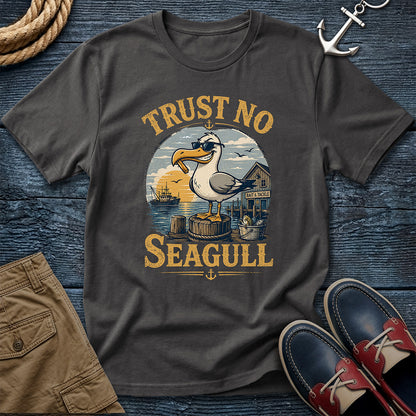 A dark charcoal gray t-shirt with a vintage-style nautical graphic of a seagull and the text 'TRUST NO SEAGULL'. The shirt is presented in a flat-lay arrangement on a dark blue wood background, styled with a rope, khaki shorts, a silver anchor, and red-and-navy boat shoes.