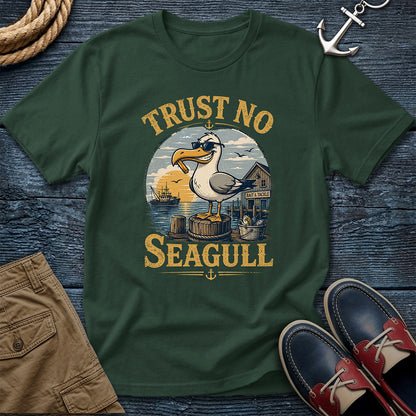 A dark green crewneck t-shirt featuring a vintage-style graphic of a seagull standing on a barrel with the text 'TRUST NO SEAGULL' in yellow. The shirt is displayed in a flat-lay arrangement on a dark wood background, surrounded by nautical-themed props including an anchor, rope, khaki shorts, and blue-and-red boat shoes.