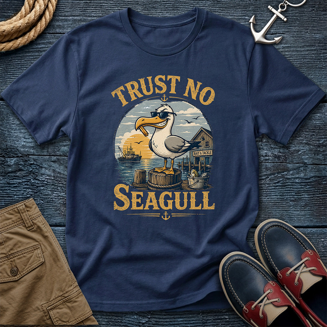 A navy blue graphic t-shirt featuring a seagull and the text 'TRUST NO SEAGULL' in yellow, styled in a nautical flat-lay with brown shorts, red and navy boat shoes, a rope, and an anchor on a dark wood background.