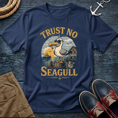 A navy blue graphic t-shirt featuring a seagull and the text 'TRUST NO SEAGULL' in yellow, styled in a nautical flat-lay with brown shorts, red and navy boat shoes, a rope, and an anchor on a dark wood background.