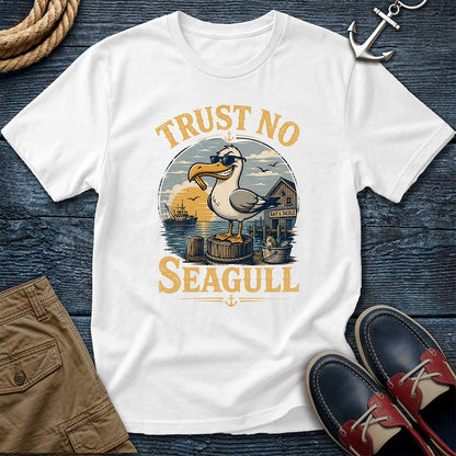 A white short-sleeve graphic T-shirt laid flat on a dark blue wood surface, surrounded by nautical styling props including a rope, anchor, khaki shorts, and navy-and-red boat shoes. The shirt graphic shows a seagull standing on a dock with the text 'TRUST NO SEAGULL' in tan lettering.
