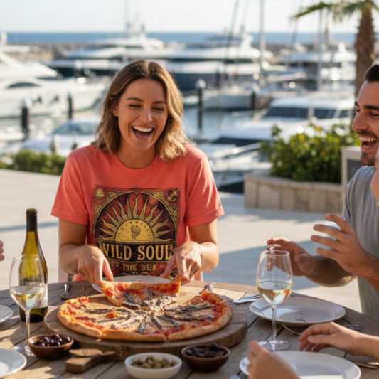 A woman wearing a coral pink graphic t-shirt with a 'Wild Soul's of the Sea' sun design sits at an outdoor wooden table with a large pizza, surrounded friends in a sunny marina setting.