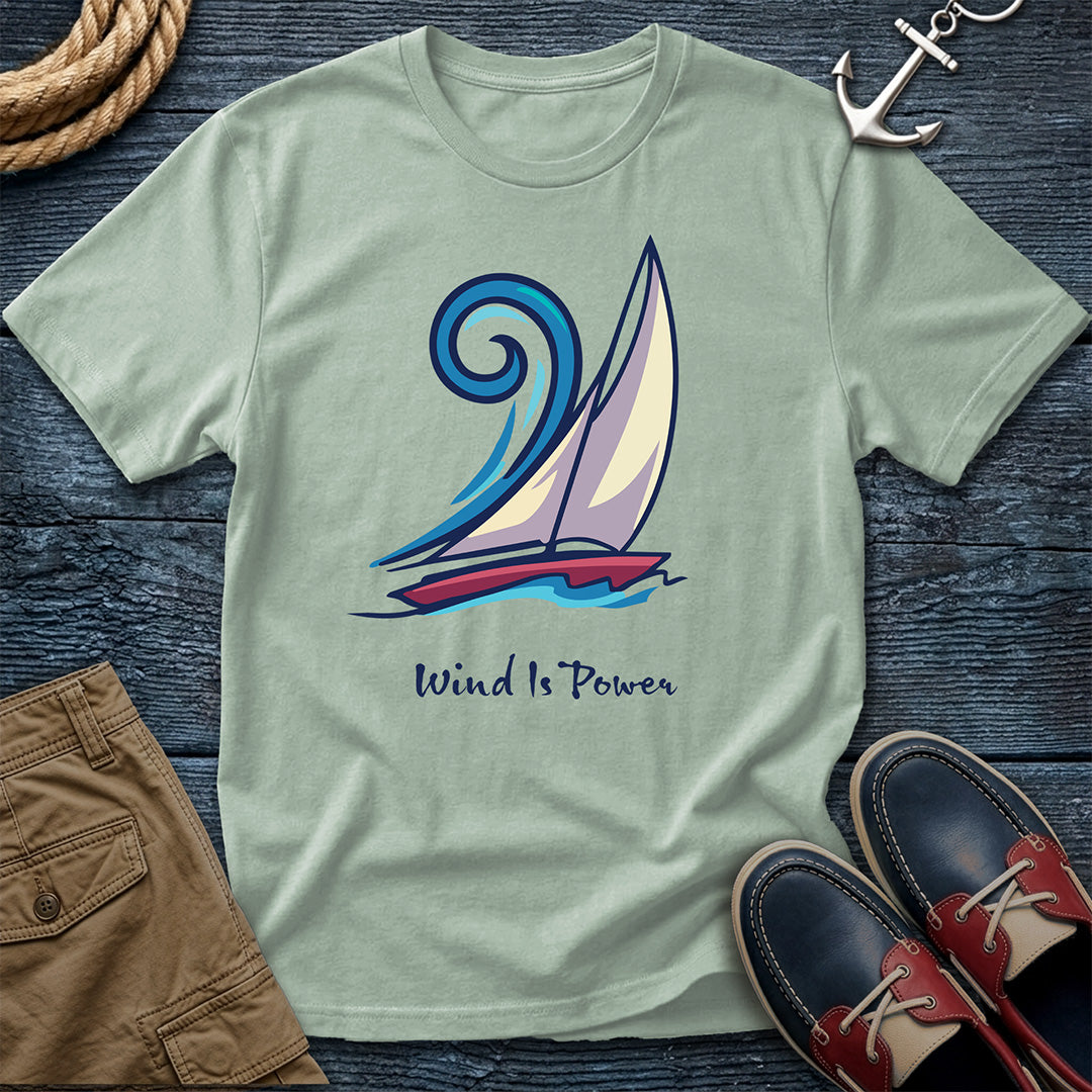 A sage green crewneck t-shirt featuring a sailboat graphic and the text 'Wind Is Power', styled in a flat lay on a dark wood background with a rope, an anchor, brown shorts, and boat shoes.