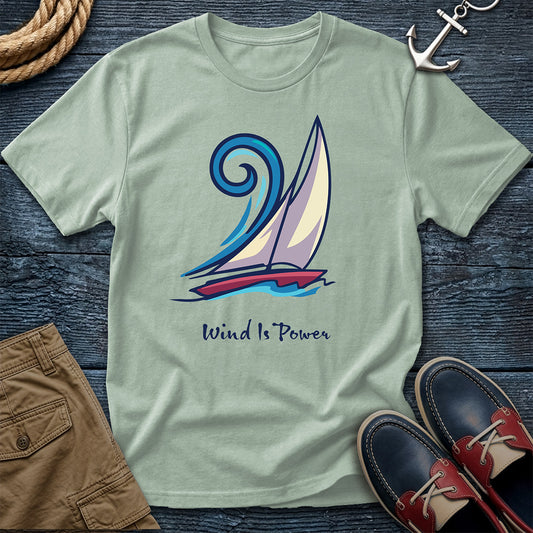 A sage green crewneck t-shirt featuring a sailboat graphic and the text 'Wind Is Power', styled in a flat lay on a dark wood background with a rope, an anchor, brown shorts, and boat shoes.
