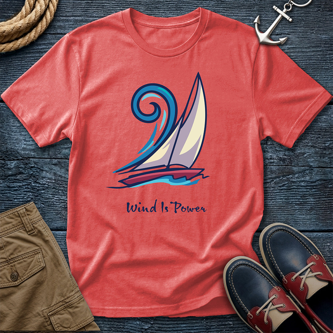 A coral red t-shirt laid flat on a dark wood background, featuring a graphic of a sailboat with the text 'Wind Is Power'. The shirt is styled with a rope, an anchor, khaki shorts, and navy blue boat shoes.