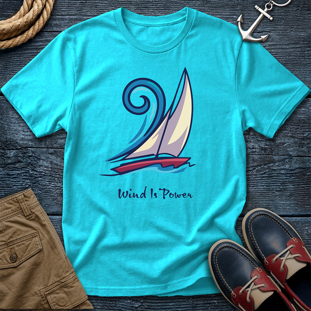 A turquoise short-sleeve graphic T-shirt laid flat on a dark wood surface, featuring a stylized sailboat illustration and the words 'Wind Is Power.' Surrounding props include a rope, an anchor, tan shorts, and navy-and-red boat shoes.