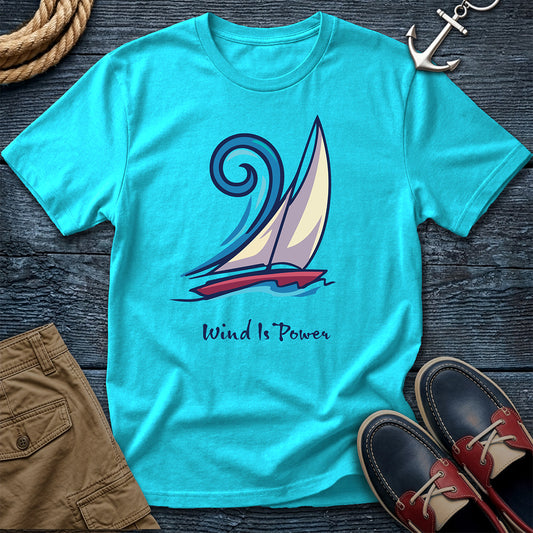 A turquoise short-sleeve graphic T-shirt laid flat on a dark wood surface, featuring a stylized sailboat illustration and the words 'Wind Is Power.' Surrounding props include a rope, an anchor, tan shorts, and navy-and-red boat shoes.
