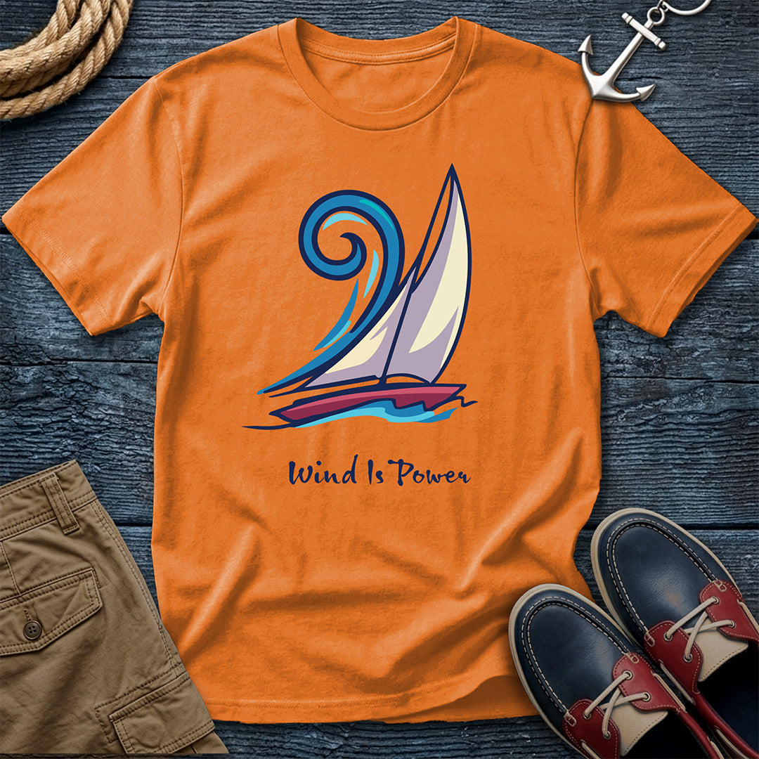An orange crewneck t-shirt featuring a sailboat graphic and the text 'Wind Is Power', styled in a flat lay on a dark wood background with a rope, khaki shorts, a silver anchor, and navy shoes.