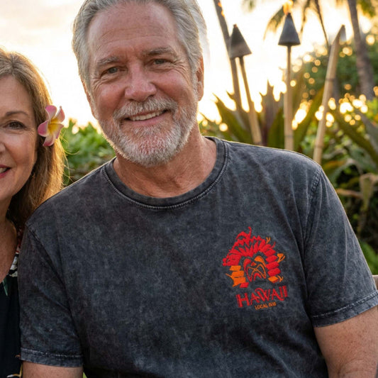 Man wearing a embroidered 'Hawaii Local' t-shirt with a woman beside him, set against a tropical background.